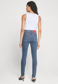 Jeans skinny en denim bleu moyen, taille haute avec une étiquette rouge "HUGO" à l'arrière. Assorti à un haut sans manches blanc et des talons noirs.
