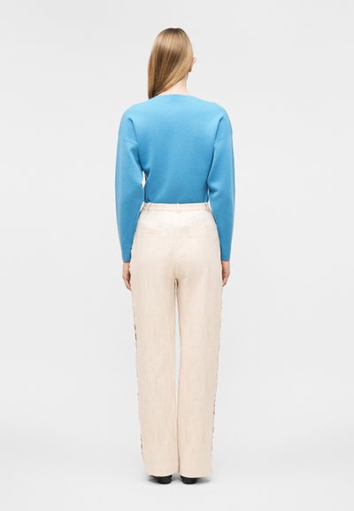 Γυναίκα με ίσια ξανθά μαλλιά που φοράει μπλε μπλούζα μακρυμάνικη και εκρού παντελόνι με φαρδιά μπατζάκια και ψηλή μέση, όρθια και γυρισμένη προς τα πίσω.