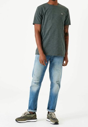 Mężczyzna w ciemnozielonej koszulce w paski z krótkim rękawem, jasnoniebieskich podniszczonych dżinsach z podwiniętymi nogawkami i oliwkowozielonych tenisówkach, stojący na białym tle.