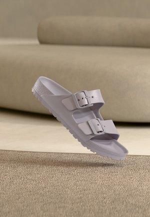 Witte sandaal met open teen en twee verstelbare gespen, zwevend boven een beige tapijtvloer en een lichte zachte achtergrond.