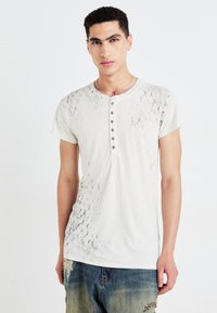 Kortärmad, ljusbeige T-shirt med ett mörkt, sprucket mönster på ena axeln, med en henley-krage och fem mörka knappar.