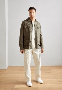 Chaqueta ligera de color verde oliva con cierre de botones y cuatro bolsillos, combinada con pantalones cargo de pierna ancha en color blanco roto y zapatillas blancas.
