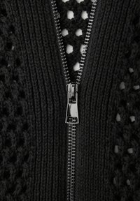 Close-up van een zwart gebreid kledingstuk met een zilveren metalen ritssluiting die gedeeltelijk open is, waarbij een gestructureerd open gebreid patroon eronder zichtbaar is.