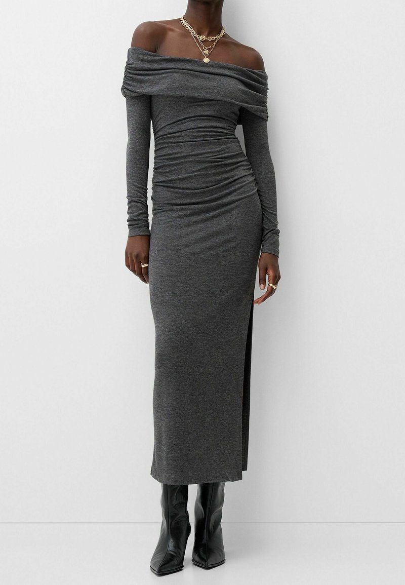 Femme portant une longue robe gris foncé épaules dénudées avec des détails froncés, des bottes noires pointues et des colliers et bagues dorés superposés.