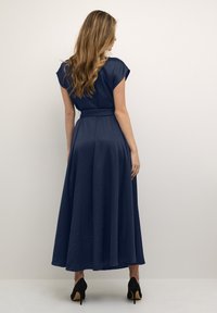 Marineblaues, bodenlanges Kleid mit kurzen Ärmeln, Taillengürtel und glatter Textur. Getragen mit spitzen schwarzen Pumps.