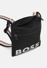 Czarna torebka na ramię z białym logo "BOSS", podwójne zamki błyskawiczne, regulowany pasek w paski w kolorze beżowym, białym i czarnym na białym tle.