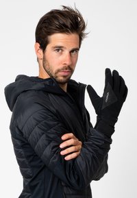 Gants noirs avec une prise texturée, arborant des motifs en diamant et un poignet sécurisé. Le modèle porte une veste noire à capuche avec des détails matelassés.