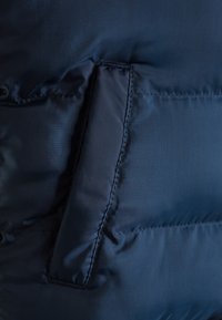 Tmavě modrá prošívaná bunda s hladkou texturou, s boční kapsou všitou do látky, zobrazená na rovné ploše.