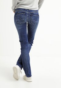 Blå denim jeans med slim fit, två bakfickor med zigzag-mönster, i kombination med vita sneakers. Slät textur och lätt stretch.