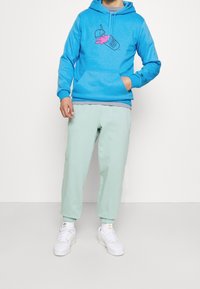 Blå huvtröja med grafiskt tryck i rosa, med en känguruficka. Kombinerad med ljusgröna joggers och vita sneakers.