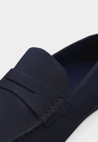 Κοντινή λήψη ενός μοκασίνι από μπλε σουέντ, με λουράκι penny και ορατή ραφή σε λευκό φόντο.