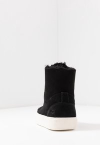 Czarne zamszowe botki UGG z futrzanym wykończeniem i białą gumową podeszwą, pokazane z tyłu na białej powierzchni i w kącie ściany.