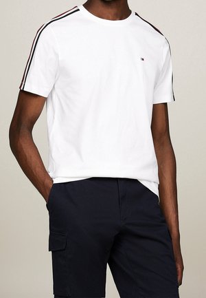 Mann trägt ein weißes kurzärmeliges T-Shirt mit roten und schwarzen Streifen auf den Schultern und marineblaue Cargoshorts, steht mit den Händen in den Taschen.