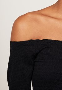 Černý top bez ramínek vyrobený z texturované tkaniny, s rovnou výstřihovou linií a dlouhými rukávy s jemně přiléhavým střihem.