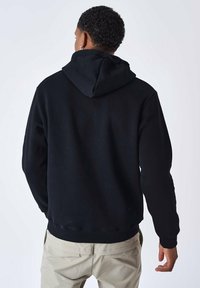 Sudadera negra con bolsillo canguro, capucha ajustable y puños de canalé. Combinada con pantalones de color claro. Textura de tela suave.