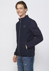 Mørkeblå pufferjakke med lynlåsfront, quiltet tekstur og kontrasterende stofærmer. Indeholder sidelommer med lynlås og et logo-detalje på brystet.