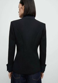 Černý oblekový blazer s strukturovaným střihem. Má klopený límec, dlouhé rukávy s manžetami a texturovanou tkaninu.