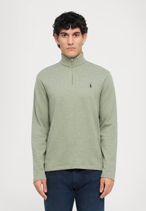 Light green zip-up sweatshirt with long sleeves, soft texture, ribbed collar, and a small navy logo on the left chest, paired with dark denim jeans.
