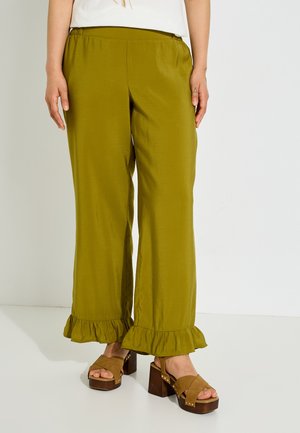 Persona que lleva pantalones de pernera ancha de color verde oliva con dobladillos con volantes y sandalias marrones de plataforma, de pie frente a un fondo blanco simple.