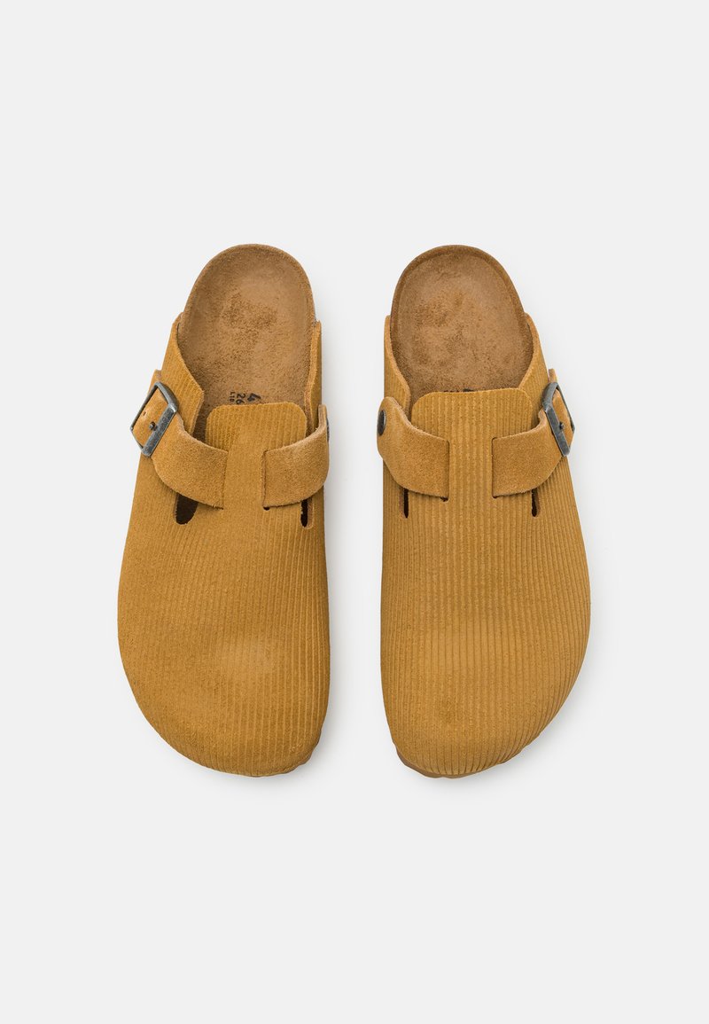 Gelbe Wildleder-Clogs mit gerillter Textur, geschlossenem Rücken, verstellbarem Riemen und offenen Seiten für Belüftung.