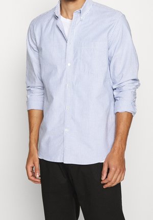 Mand iført en lyseblå og hvid vertikalt stribet skjorte med opklappede ærmer og sorte bukser.