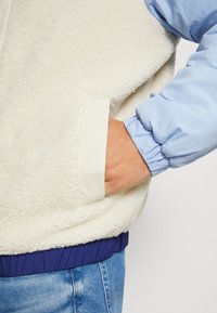 Giacca di colore crema foderata in pile con uno strato esterno blu e polsini elasticizzati, caratterizzata da una rifinitura delle tasche in crema e un orlo a coste blu scuro.