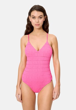 Joven con cabello largo y castaño que lleva un bañador rosa texturizado de una sola pieza con tirantes delgados, posando frente a un fondo claro y liso.