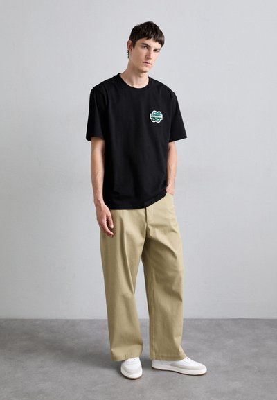 Black cotton t-shirt with a green embroidered logo on the chest, paired with light beige wide-leg pants and white sneakers.