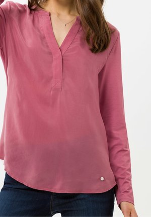 Femme portant un chemisier rose poudré à manches longues avec un col en V et un jean bleu foncé sur un fond uni.