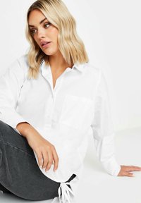 Camisa blanca de botones con un único bolsillo en el pecho, mangas largas y un corte holgado. La tela parece suave y ligera.