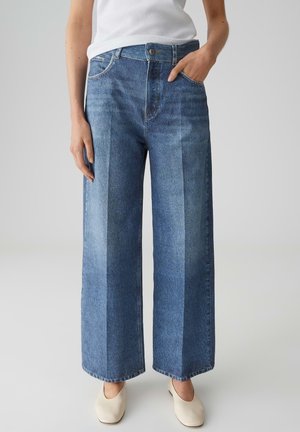 Vrouw met een witte top, crème instapschoenen en een high-waist wijde blauwe jeans met zichtbare naden aan de voorkant.
