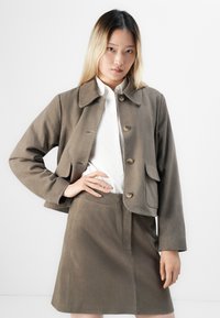 Mladá žena s dlouhými ombre vlasy má na sobě taupe semišovou bundu a odpovídající áčkovou sukni přes bílou košili s knoflíky, stojí proti bílé stěně.
