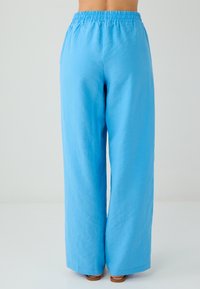 Persona che indossa pantaloni blu larghi a vita alta con elastico in vita, in piedi a piedi nudi su uno sfondo chiaro e semplice, vista da dietro.