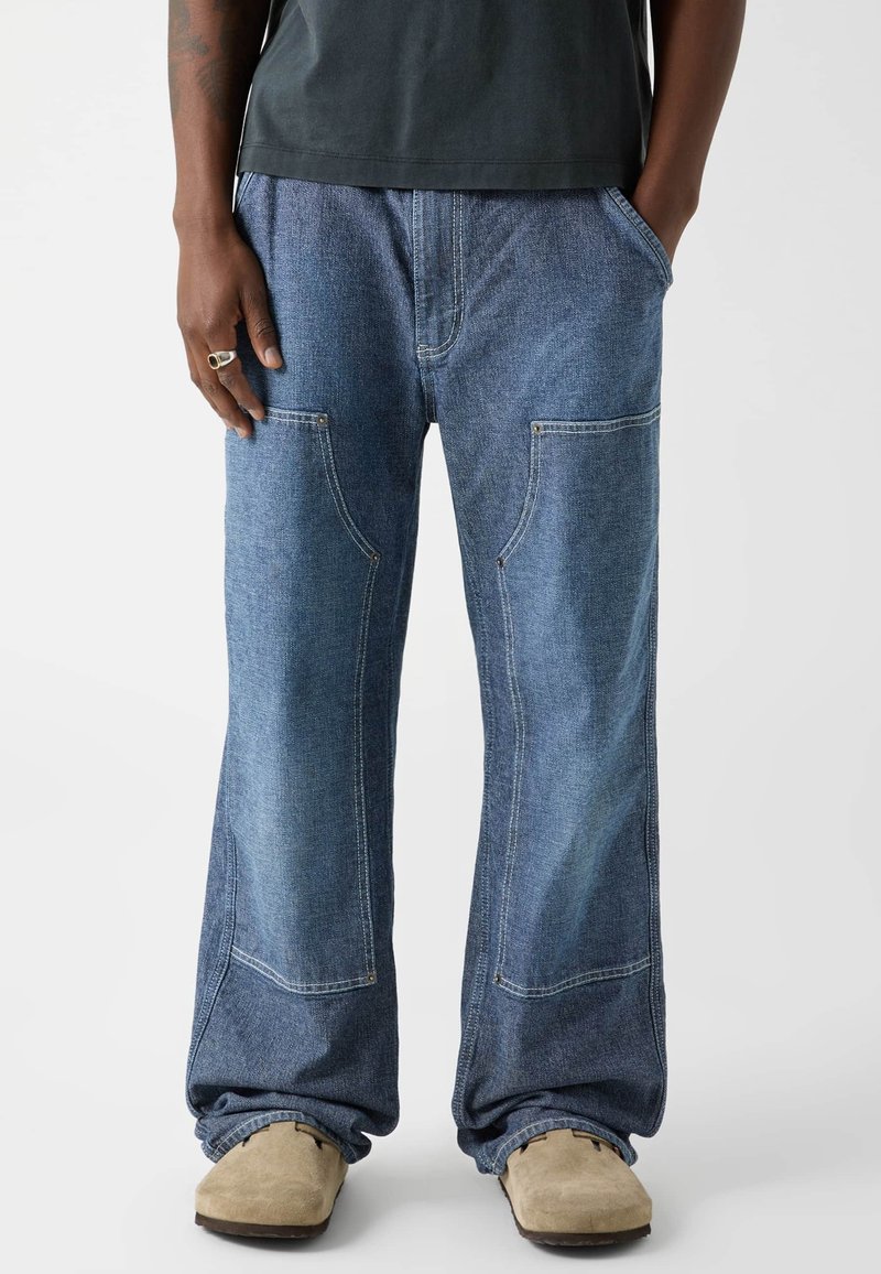 Personne portant un jean bleu ample avec des coutures distinctives et des chaussures beiges à enfiler, debout les mains dans les poches.