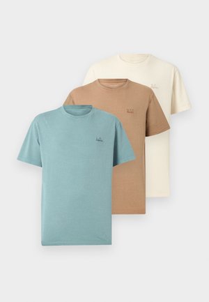 Homme portant une casquette marron, un t-shirt vert clair, un jean bleu clair et des chaussures marron, ajustant la casquette avec une expression souriante.