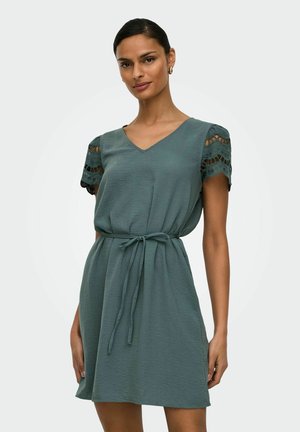 Ženska v turkizni obleki s kratkimi rokavi z čipkastimi izrezi na rokavih in pasom zvezanim v vozel, stoji pred enobarvnim ozadjem.