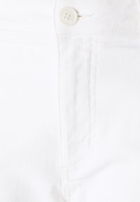 Tejido de mezclilla blanca con un bolsillo lateral, un botón visible y una textura sutil. Los detalles de costura a lo largo de los bordes complementan el diseño.