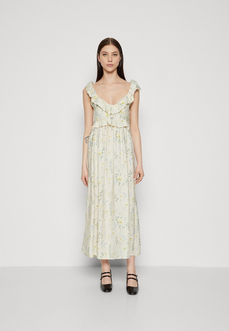 Robe maxi florale avec un col volants, fond jaune clair, motifs floraux verts et bleus, et taille cintrée. Associée à des chaussures noires.