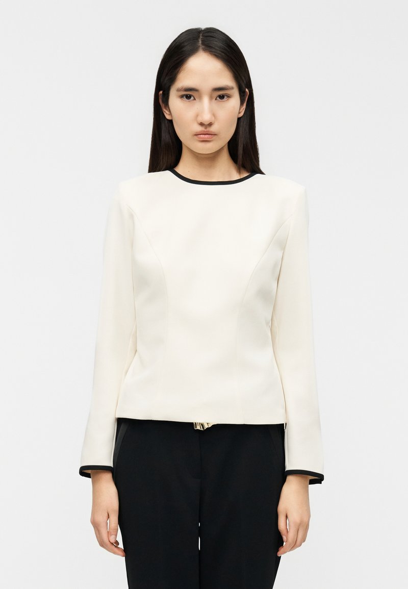 Άσπρο τοπ με δομημένη υφή και μακριά μανίκια, με μαύρο φινίρισμα στη λαιμόκοψη και τις μανσέτες, χαρακτηρίζεται από λεία υφή και εφαρμοστή σιλουέτα.