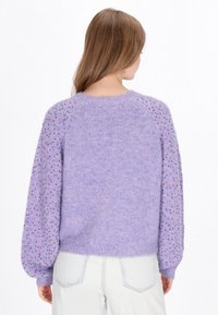 Lila Pullover mit strukturierten Raglanärmeln, die mit pinken und lila Punkten verziert sind, aus weichem Material, mit geripptem Halsbündchen und Saum, getragen mit hellblauen Jeans.