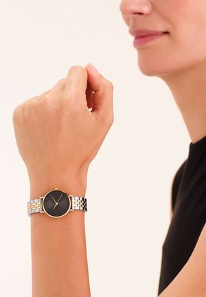 Orologio tondo nero con dettagli dorati, bracciale in metallo argento e oro, esposto su un polso su uno sfondo neutro.