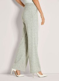 Vrouw in grijze, textuurrijke wijde broek en witte hoge hakken, staand tegen een lichte muur, zijwaarts kijkend.