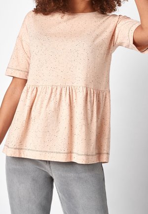 Osoba nosząca jasno różowy, nakrapiany T-shirt z baskinką i krótkimi rękawami oraz szare dżinsy, skrócone powyżej talii.