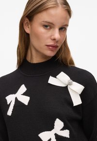 Czarny sweter z wysokim kołnierzem, ozdobiony białymi dzianymi kokardkami i wysadzaną klejnotami broszką na jednym ramieniu. Gładka faktura i dopasowany krój.