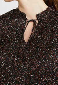 Svart blus med hög hals och volangdetaljer, knytning och ett mönster av flerfärgade stjärnor i orange, rosa och vitt.