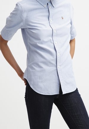 Osoba ubrana w jasnoniebieską koszulę zapinaną na guziki z małym logo na piersi oraz ciemne dopasowane dżinsy, stojąca z rękami za plecami.