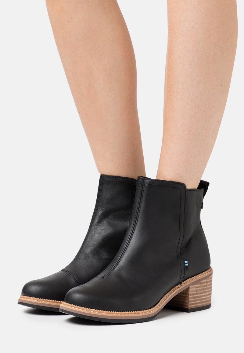 Bottes chevilles en cuir noir avec talons en bois empilés, portées sur des jambes nues contre un fond clair uni.