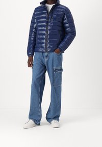 Marineblå dunjakke med vandret quiltet mønster, lynlås og opretstående krave; kombineret med løstsiddende lyseblå jeans og hvide sneakers.