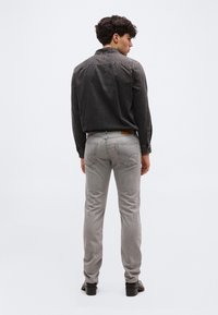 Νεαρός άνδρας με σγουρά μαλλιά, ντυμένος με μαύρο τζιν πουκάμισο και ανοιχτό γκρι τζιν, στέκεται με την πλάτη σε έναν απλό φόντο.