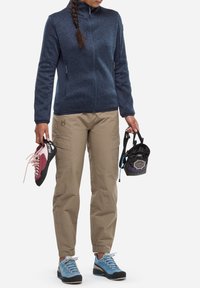 Veste en polaire navy avec des tissus lisses contrastants, pantalon cargo beige, chaussures de randonnée bleu et noir, tenant des chaussures d'escalade roses et un sac noir.